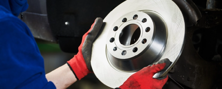 A technician in a blue boiler suit wearing red gloves holding a cars brake disc.